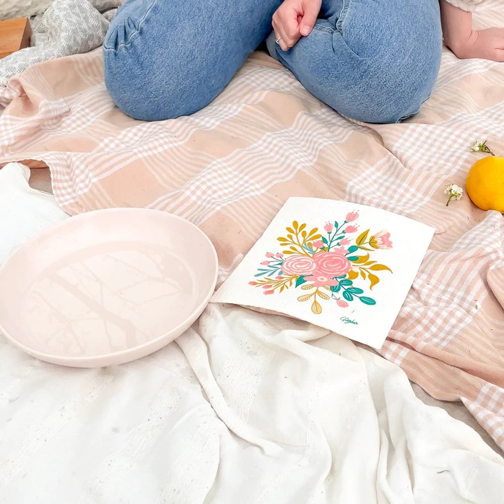 A woman is next to an empty plate and a floral bouquet patterned Goldilocks dishcloth, in a picnic setting.